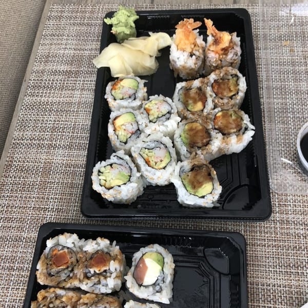 Two black takeout containers filled with various sushi rolls, ginger, and wasabi on a woven placemat.
