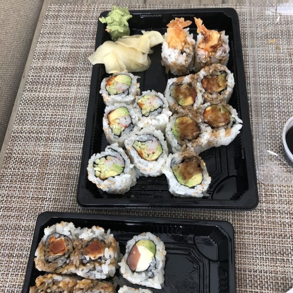 Two black takeout containers filled with various sushi rolls, ginger, and wasabi on a woven placemat.