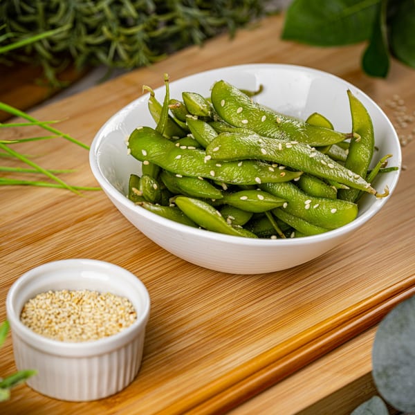 A white bowl of green edamame with sesame seeds, next to a small dish of sesame seeds.
